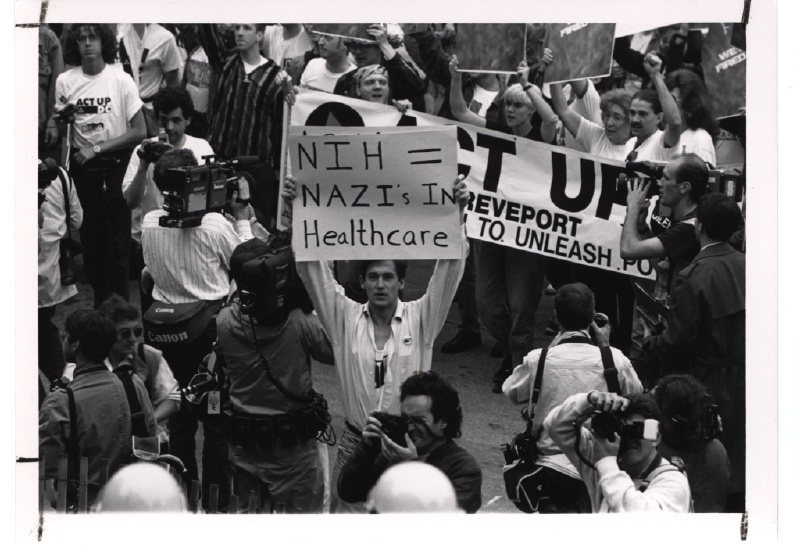 मई 1990 में अमेरिकी राष्ट्रीय स्वास्थ्य संस्थान (NIH) के समक्ष प्रदर्शन करते ACT UP आंदोलन के कार्यकर्ता। फोटो: [NIH History Office / Wikimedia Commons](https://commons.wikimedia.org/wiki/File:ACT_UP_Activists_at_the_National_Institutes_of_Health_(14359361475).jpg) (पब्लिक डोमेन)