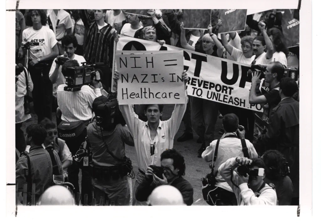 मई 1990 में अमेरिकी राष्ट्रीय स्वास्थ्य संस्थान (NIH) के समक्ष प्रदर्शन करते ACT UP आंदोलन के कार्यकर्ता। फोटो: [NIH History Office / Wikimedia Commons](https://commons.wikimedia.org/wiki/File:ACT_UP_Activists_at_the_National_Institutes_of_Health_(14359361475).jpg) (पब्लिक डोमेन)