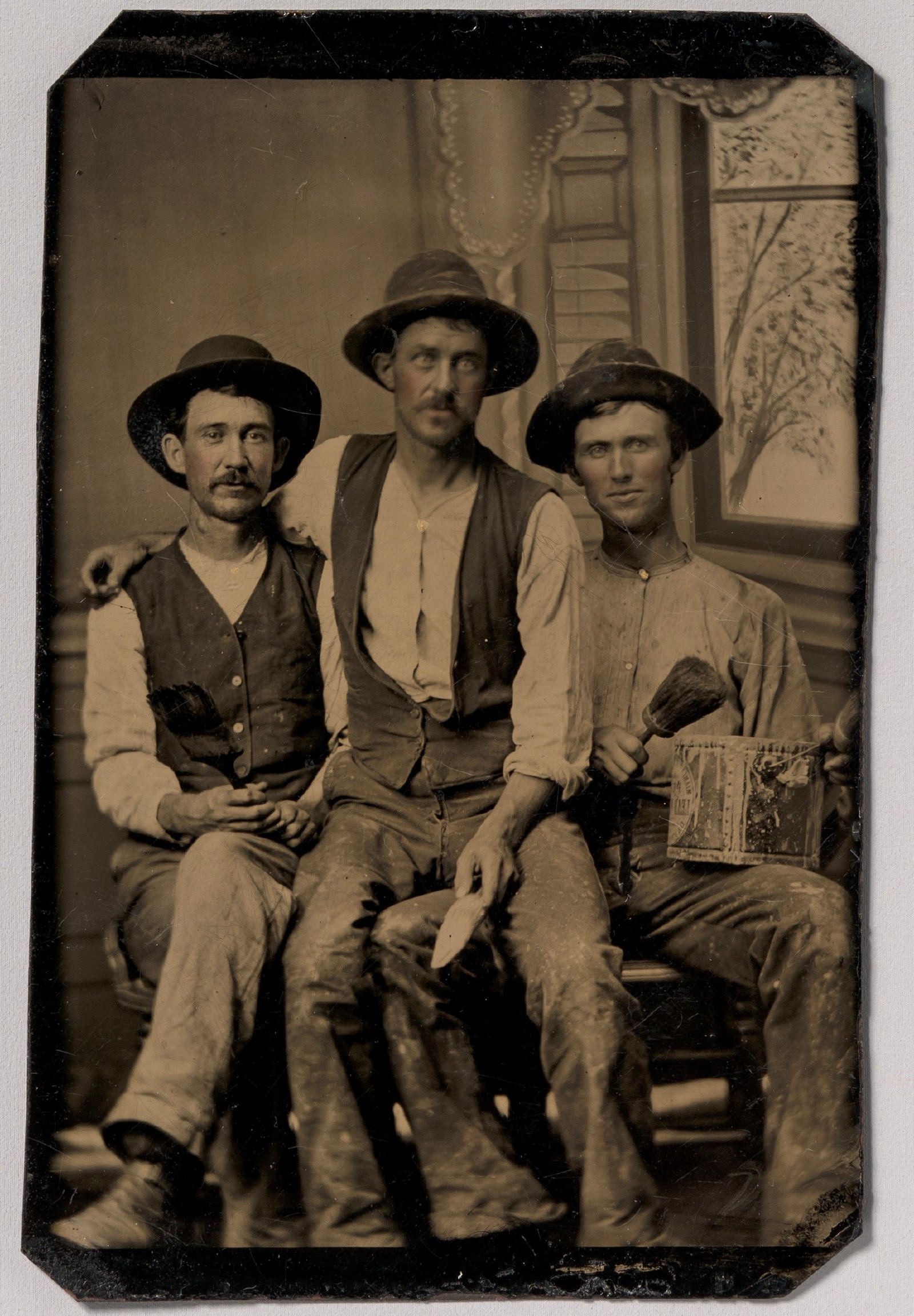 Três pintores com pincéis e uma lata de tinta diante de um cenário pintado com uma janela; ferrotipia de autor desconhecido, décadas de 1870–1880