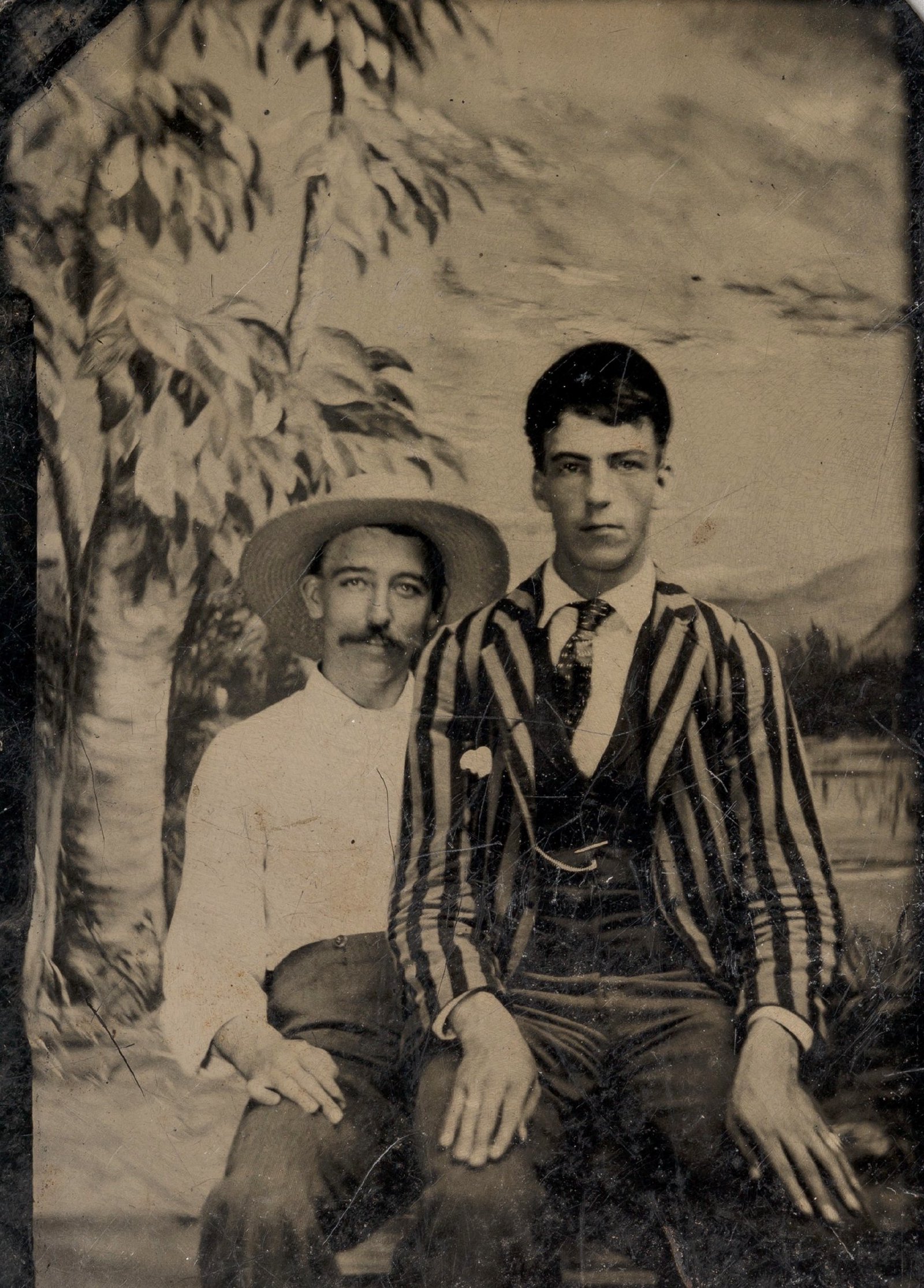 Jovem de casaco às riscas sentado no colo de outro homem diante de um cenário pintado representando uma rua; ferrotipia de autor desconhecido, décadas de 1860–1880